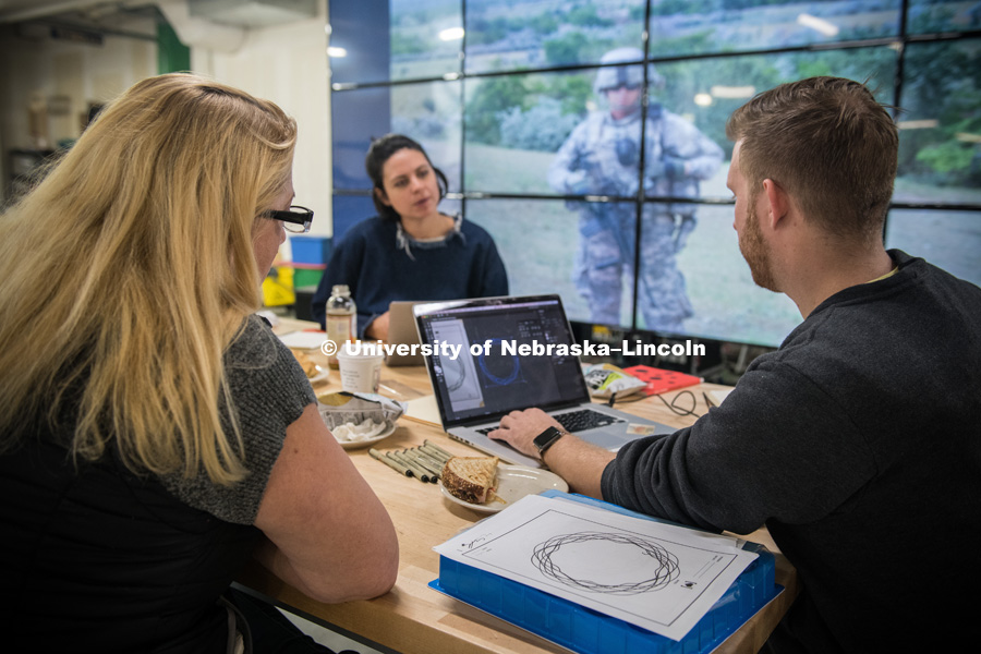 Veteran Melissa Ewing (left), Kendra Clapp Olguin, Has Heart communications director, and Husker alum Jordan Lambrecht work on the artwork representing Ewing's life story. Behind them, on screens at Nebraska Innovation Studio, is a photograph from Ewing's tour in Afghanistan. October 12, 2018. Photo by Greg Nathan, University Communication.
