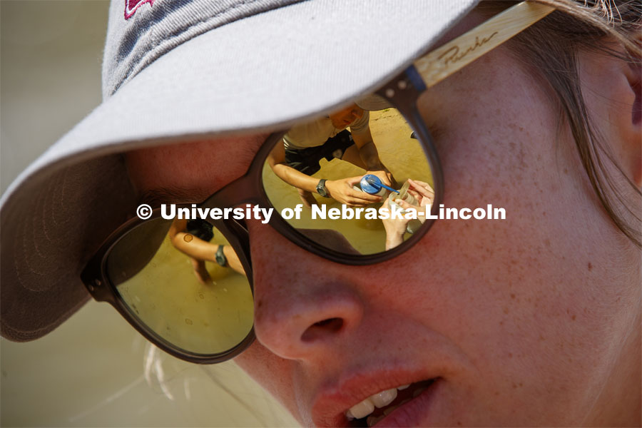 A sand sample being rinsed into a collection bottle is reflected in Kayla Vondracek's sunglasses. Jessica Corman, assistant professor in the School of Natural Resources, UCARE research group researching algae in the Niobrara River. Fort Niobrara National Wildlife Refuge. July 12, 2019. Photo by Craig Chandler / University Communication.