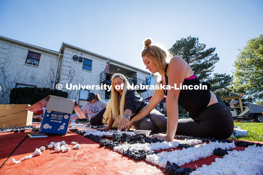 Elisa Fricke, right, freshman from Omaha, and Ainsley Rickertsen, freshman from Gothenburg, glue poms to a plywood display outside the Theta Xi house. Students from Theta Xi, Alpha Phi, Kappa Alpha Theta, Phi Kappa Theta work on their homecoming display. 2019 Homecoming display decorations. October 3, 2019. Photo by Craig Chandler / University Communication.