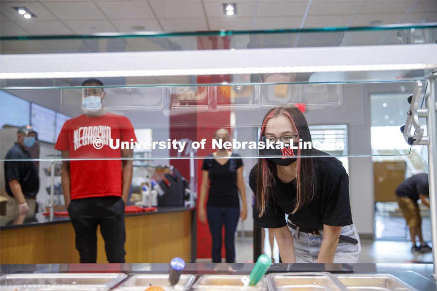 A young woman leans in toward the food bar to select her lunch at the Willa Cather Dining Center. Photo shoot of students wearing masks and practicing social distancing in dining services in Willa Cather Dining Center. July 1, 2020. Photo by Craig Chandler / University Communication.