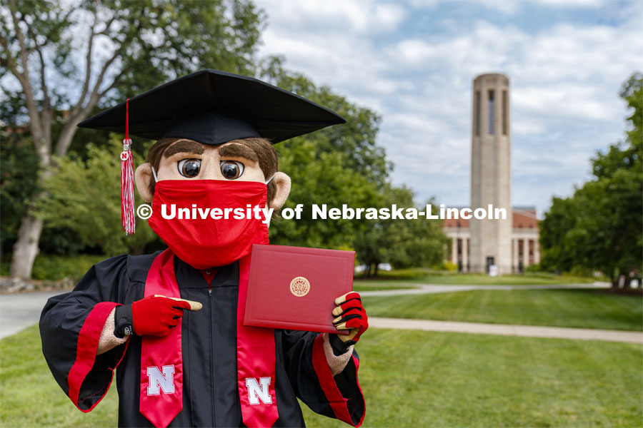 Herbie Husker has a protective mask on while he poses in front of the Mueller Bell tower while wearing a graduation cap and gown. On campus. August 5, 2020. Photo by Craig Chandler / University Communication.