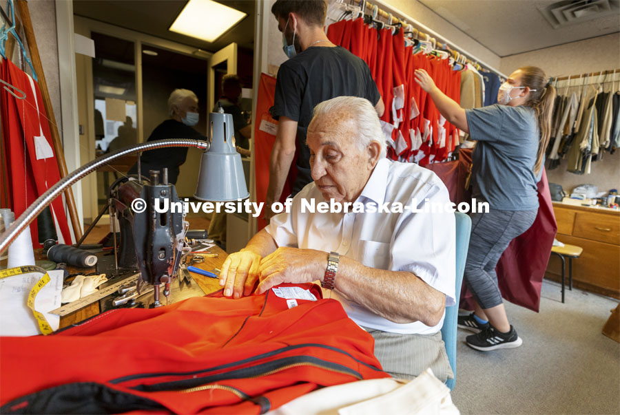 Demetrios Deligiannis (front) alters a pair of band pants as his wife Stamatia (back, left) helps band members pick up their finished uniforms before the first home game this Saturday. This year, each member of the band received new pants for the season. Each of the 300 pairs was custom tailored by the couple. Demetrios and Stamatia have been altering the Cornhusker Marching Band uniforms for 50 years from their Lincoln tailoring business. September 1, 2021. Photo by Craig Chandler / University Communication.