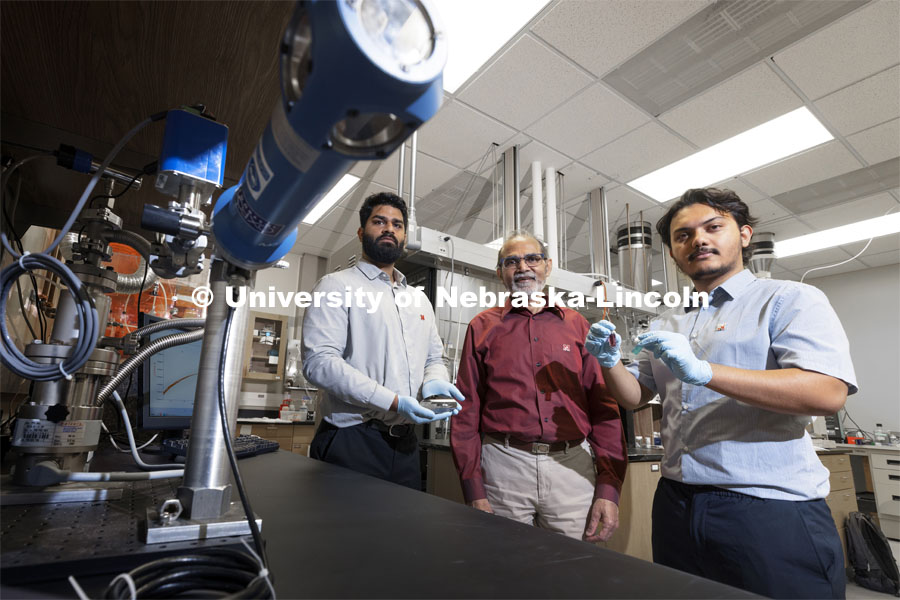 Abhijeet Prasad, a PhD student in engineering, Ravi Saraf, Professor in Chemical and Biomolecular Engineering, and Aashish Subedi, an undergraduate student in Physics, are photographed in Saraf’s Othmer Hall lab. Prasad is holding a bunch of small “electronic chips” to study these “nanoparticle necklace network” devices made in the Nebraska Center for Materials and Nanoscience. Subedi is holding a suspension of nanoparticle necklaces (blue container) made from individual particles (red container). September 24, 2021. Photo by Craig Chandler / University Communication.
