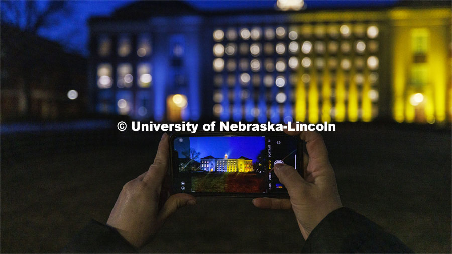 Melissa Wilkerson photographs Love Library on Nebraska’s City Campus. The library was illuminated in blue and yellow lights to show the world our hearts go out to our Husker family at home and abroad who are affected by the ongoing crisis in Ukraine. March 7, 2022. Photo by Craig Chandler / University Communication.