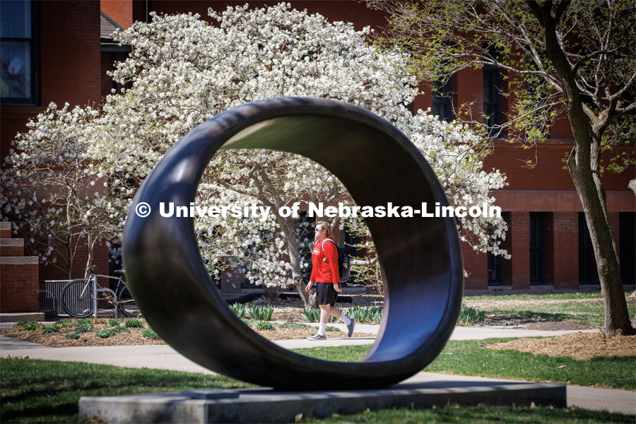 A student crossing campus in front of Architecture Hall is framed by the Fragment XO sculpture. City Campus. April 13, 2023. Photo by Craig Chandler / University Communication.
