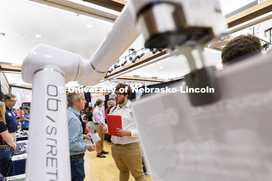 Framed by a robotic arm, Leandro Castellanos Izaguirre, a mechanical engineering and physics major, talks with representatives of CED at the Career Fair in the Nebraska Union. September 20, 2023. Photo by Craig Chandler / University Communication.