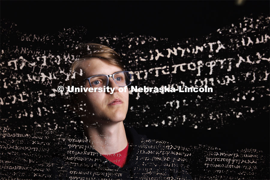 Luke Farritor, a senior at Nebraska in the Jeffrey S. Raikes School of Computer Science and Engineering, with superimposed Greek text from a nearly 2,000-year-old scroll that his work is helping to decipher. Farritor, recently won a global contest to read the first text inside a carbonized scroll from the ancient Roman city of Herculaneum. That text had been unreadable since the eruption of Mount Vesuvius in A.D. 79. Farritor developed a machine-learning algorithm that detected the Greek word πορϕυρας (purple) on the charred papyrus scroll, which is too delicate to unroll using MRI scans. In this multiple exposure, the word πορϕυρας is in the line of text appearing in the same plane to the right of his eyes. November 3, 2023. Photo by Craig Chandler / University Communication and Marketing.