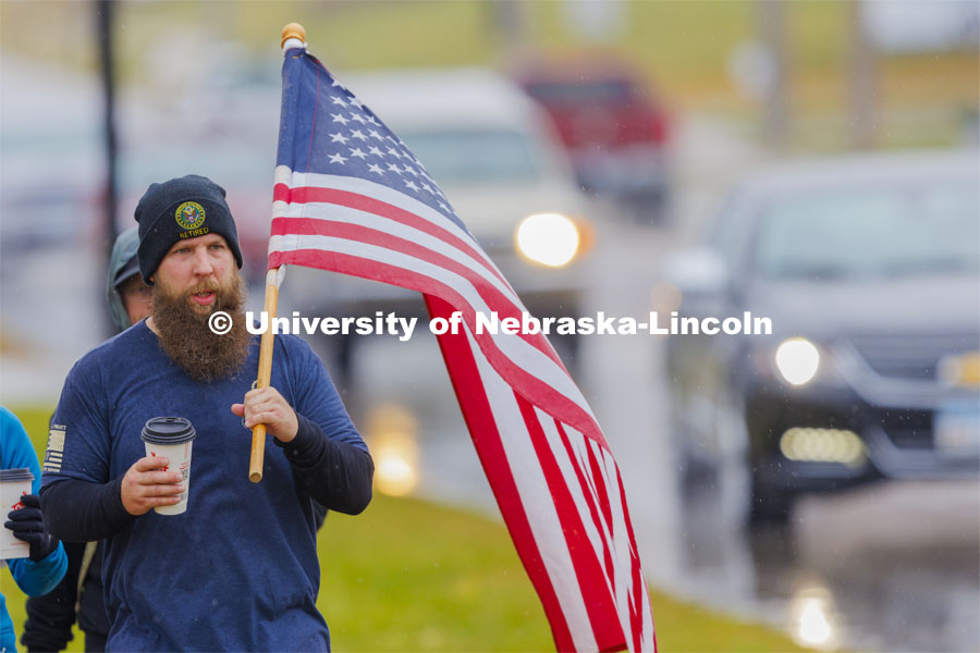 Army veteran and Iowa resident Casey Swanson carries the flag as they walk into Atlantic, Iowa. Employees from a convience store brought out coffee for the marchers. Ruck Marchers walk along Hwy 83 east of Atlantic, Iowa, carrying the Nebraska v. Iowa game ball. The Things They Carry" ruck march involving military and veteran students from Iowa and Nebraska. To raise awareness about veteran suicide, through the week, the students walk in 20-mile shifts carrying 20-pound backpacks to commemorate the estimated 20 veterans who die by suicide each day. November 20, 2023. Photo by Craig Chandler / University Communication and Marketing