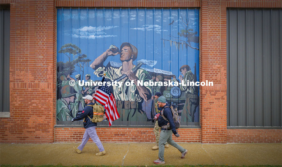 Nebraska alumni and veteran Pete Lass carries the American flag as he and Atlantic-area veterans Trevor Gipple (carrying game ball) and Ryan Graham walk past a mural on the side of the Coca-Cola Atlantic Bottling Company building in downtown Atlantic, Iowa, on the second day of the march. Ruck Marchers walk along Hwy 83 east of Atlantic, Iowa, carrying the Nebraska v. Iowa game ball. The Things They Carry" ruck march involving military and veteran students from Iowa and Nebraska. To raise awareness about veteran suicide, through the week, the students walk in 20-mile shifts carrying 20-pound backpacks to commemorate the estimated 20 veterans who die by suicide each day. November 20, 2023. Photo by Craig Chandler / University Communication and Marketing.