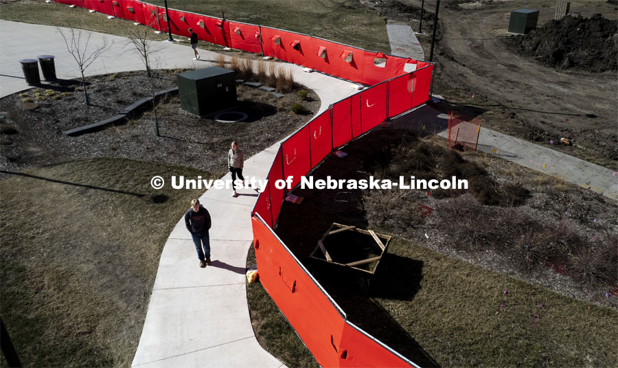 Students cross campus. A red fence surrounds the construction area on East Campus Legacy Plaza in the greenspace. February 26, 2024. Photo by Craig Chandler / University Communication and Marketing.