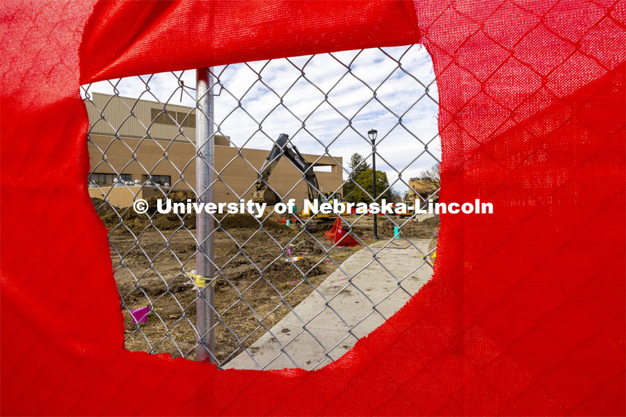 A hole in the construction fence frames the backhoe at work. A red fence surrounds the construction area on East Campus Legacy Plaza in the greenspace. February 26, 2024. Photo by Craig Chandler / University Communication and Marketing.