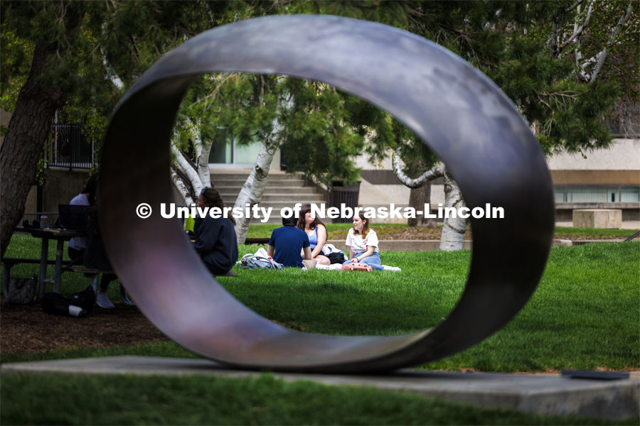 Framed by the sculpture Fragment X-O, Paige Vose, center, Makena Niehaus and Evan Mott (back to camera) enjoy the sunshine and awesome weather at the Sheldon sculpture garden. April 17, 2024. Photo by Craig Chandler / University Communication and Marketing.