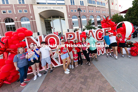 Several families of Husker fans pose in front of a sign that reads "No Place Like Home (Nebraska)" d