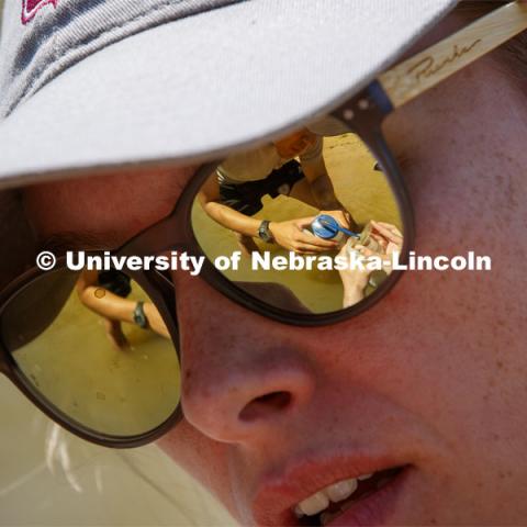 A sand sample being rinsed into a collection bottle is reflected in Kayla Vondracek's sunglasses. Jessica Corman, assistant professor in the School of Natural Resources, UCARE research group researching algae in the Niobrara River. Fort Niobrara National Wildlife Refuge. July 12, 2019. Photo by Craig Chandler / University Communication.