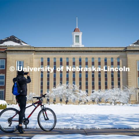 A young man riding by stops to take a picture of Love Library. An April snowstorm leaves campus unseasonably beautiful. April 17, 2020. Photo by Craig Chandler / University Communication.