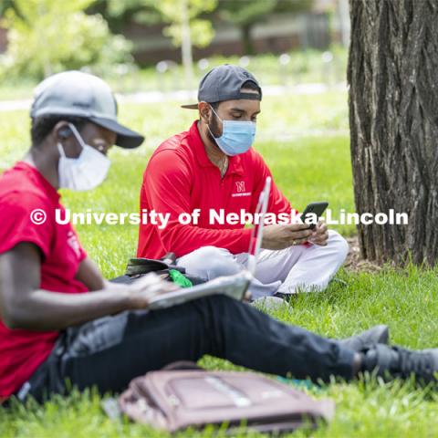 Mark Iradukunda works on his laptop and Aldi Airori checks his phone while wearing masks and practicing social distancing outdoors. Photo shoot of students wearing masks and practicing social distancing. June 24, 2020. Photo by Craig Chandler / University Communication.