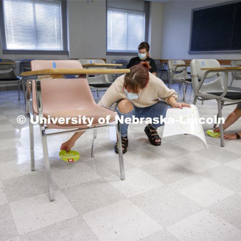 Student worker Macy Behrens, left, and Justy Bullington with the Executive Vice Chancellor's office, place seat-available stickers on the floor under a desk in a Louise Pound Hall classroom.  The stickers allow the seats to be moved by the janitorial crew for cleaning and be placed back in the correct spots. Crews mark classroom seats in Louise Pound Hall as part of preparing the campus for Forward to Fall. A crew is going room-to-room marking the where to sit and where the chairs/tables go. By Friday, they hope to have 483 classrooms and 8970 seats on all campuses marked and ready. August 3, 2020. Photo by Craig Chandler / University Communication.