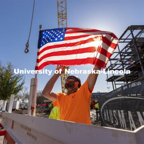 A Hausmann Construction employee unfurls a flag placed on the final steel beam during the topping out ceremony on April 9. The ceremony has roots to an ancient Scandinavian tradition that, in modern times, signals the completion of the internal structure of a building. Topping out ceremony for the new construction atop the demolished Mabel Lee Hall. April 9, 2021. Photo by Craig Chandler / University Communication.