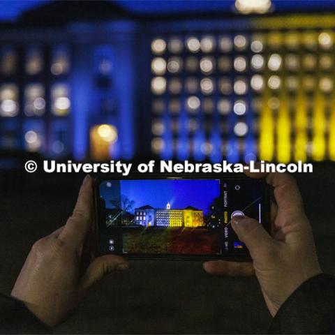 Melissa Wilkerson photographs Love Library on Nebraska’s City Campus. The library was illuminated in blue and yellow lights to show the world our hearts go out to our Husker family at home and abroad who are affected by the ongoing crisis in Ukraine. March 7, 2022. Photo by Craig Chandler / University Communication.