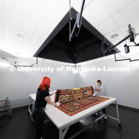 Student workers Olivia Fiore, left, and Inas Hskan, fold a quilt after it was photographed.  The quilt is titled “Soldier’s Quilt” c. 1850-1910 and probably made in India. The area above the quilt leads to a second-floor balcony with a camera suspended above the table for photographs. Quilt Center. February 8, 2023. Photo by Craig Chandler / University Communication.