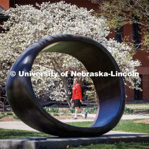 A student crossing campus in front of Architecture Hall is framed by the Fragment XO sculpture. City Campus. April 13, 2023. Photo by Craig Chandler / University Communication.
