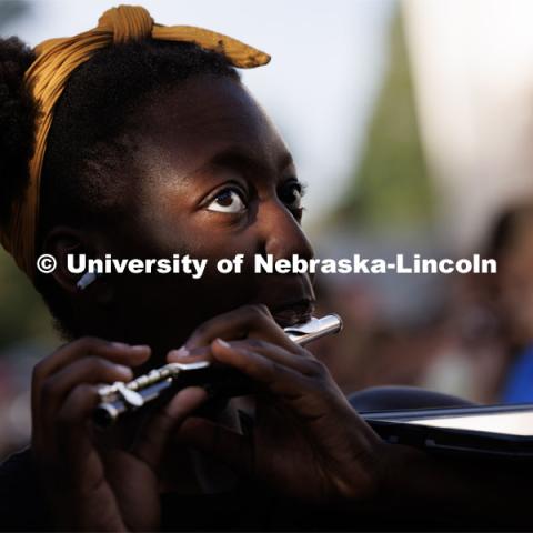 Tori Thomas watches Tony Falcone direct the band as the sun dapples her face through the trees. The band practices on the greenspace by the Sheldon Art Museum as their normal pregame concert area next to the Kimball Recital Hall is under construction. Cornhusker Marching Band practice. August 16, 2023. Photo by Craig Chandler/ University Communication.