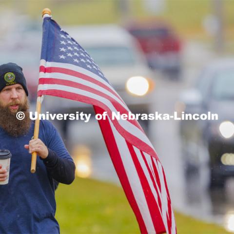 Army veteran and Iowa resident Casey Swanson carries the flag as they walk into Atlantic, Iowa. Employees from a convience store brought out coffee for the marchers. Ruck Marchers walk along Hwy 83 east of Atlantic, Iowa, carrying the Nebraska v. Iowa game ball. The Things They Carry" ruck march involving military and veteran students from Iowa and Nebraska. To raise awareness about veteran suicide, through the week, the students walk in 20-mile shifts carrying 20-pound backpacks to commemorate the estimated 20 veterans who die by suicide each day. November 20, 2023. Photo by Craig Chandler / University Communication and Marketing