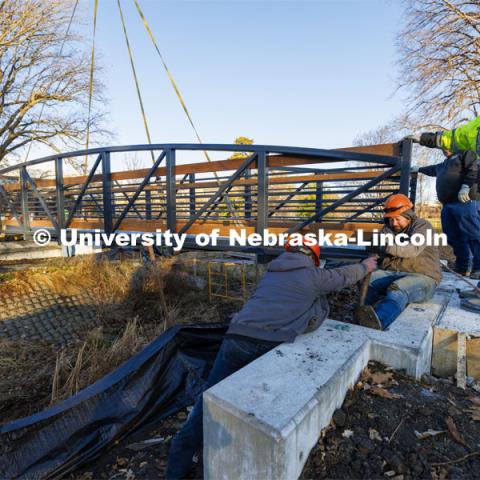 Workers with Judds Brothers Construction adjust a long bolt so it aligns with the bridge as it is lowered onto the bolts on the east side of the creek. A new footbridge over Dead Mans Run on East Campus’ Maxwell Arboretum. December 5, 2023. Photo by Craig Chandler / University Communication and Marketing.