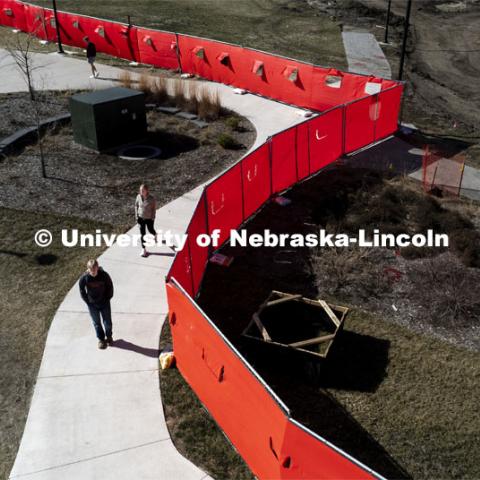 Students cross campus. A red fence surrounds the construction area on East Campus Legacy Plaza in the greenspace. February 26, 2024. Photo by Craig Chandler / University Communication and Marketing.