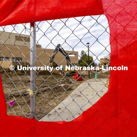 A hole in the construction fence frames the backhoe at work. A red fence surrounds the construction area on East Campus Legacy Plaza in the greenspace. February 26, 2024. Photo by Craig Chandler / University Communication and Marketing.