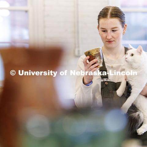 Kat Kastens, junior, looks at a cup while holding her cat Leo during the UNL Clay Club sale inside Richards Hall. December 13, 2024. Photo by Jordan Opp / University Communication and Marketing